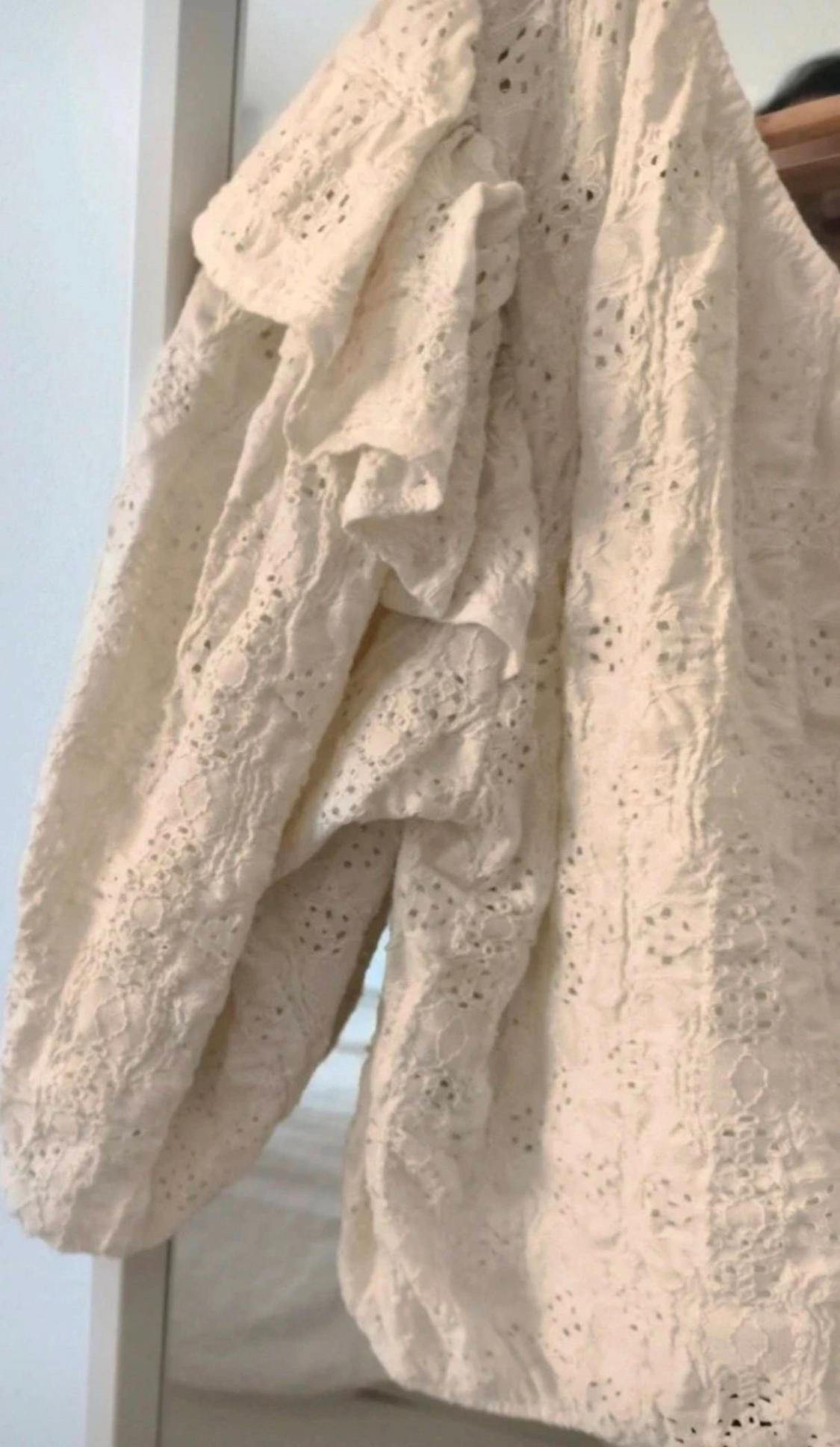 Close-up of a white garment with broderie anglaise detailing and ruffles, hanging on a wooden hanger in a bright, relaxed setting.