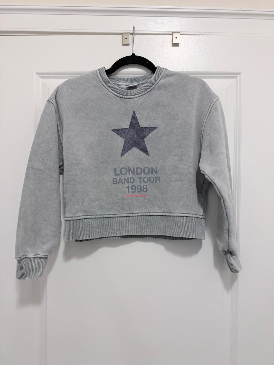 Gray long-sleeve sweatshirt hanging on a white door. It features a star and the text LONDON BAND TOUR 1998.