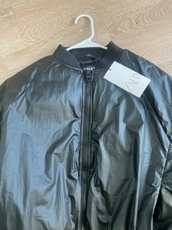 Black jacket with zipper and round neck on a white hanger against a light wood background. Visible label with text "ZARA".