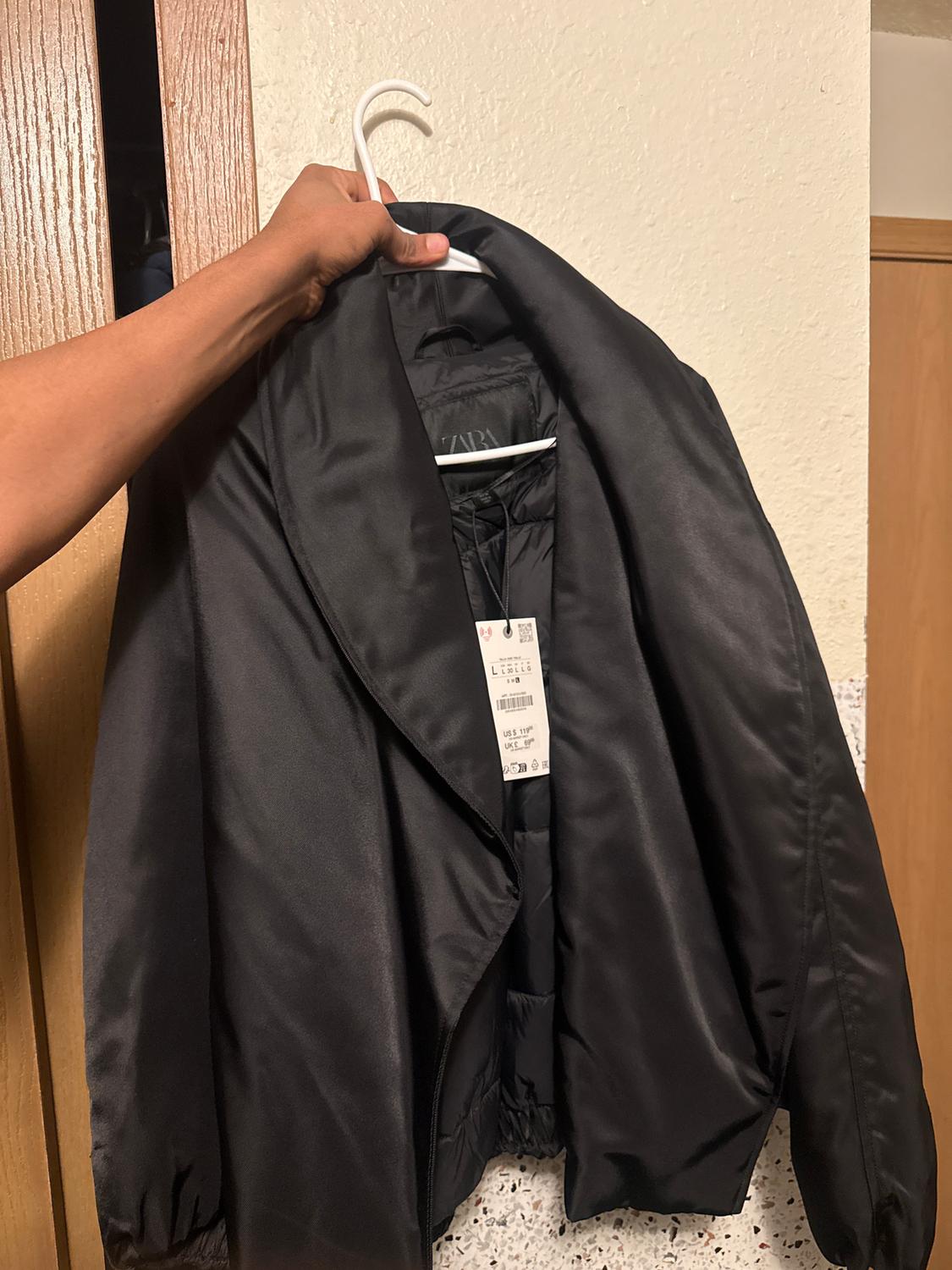 A black jacket is hanging on a white hanger, held by a hand. The label is visible on the inside. The background is a light-coloured wall with a wooden door.