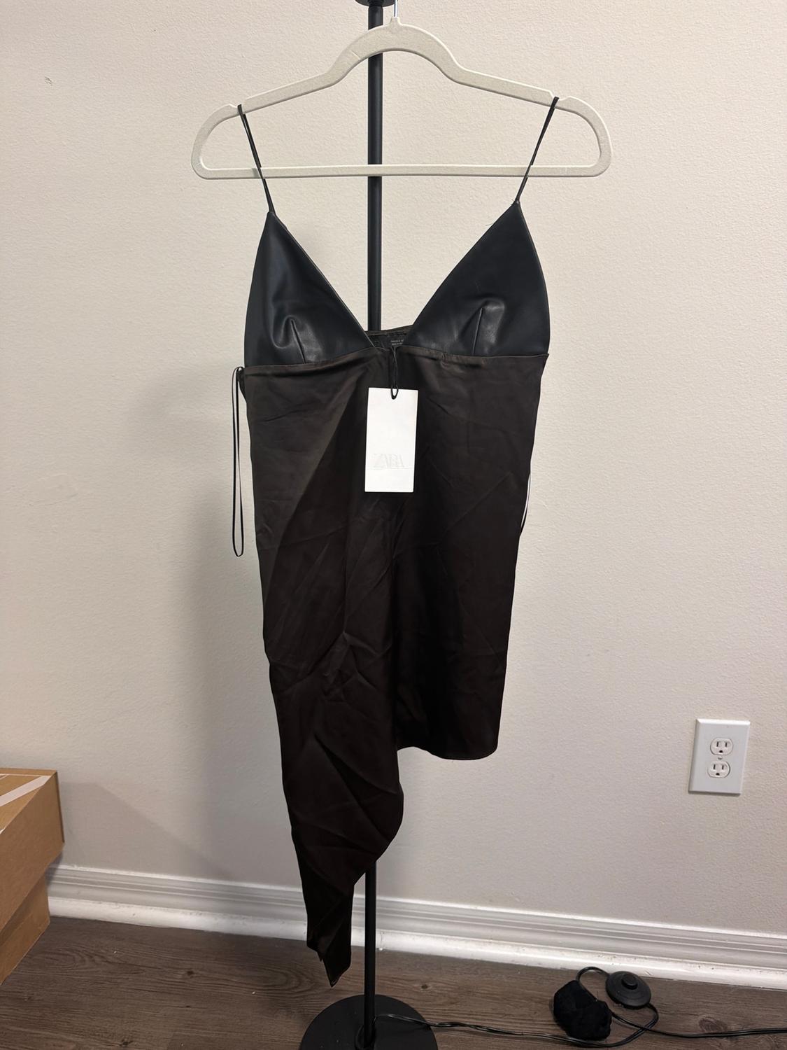 Asymmetrical black dress with thin straps on a hanger, visible tag. White background, wooden floor, and cardboard box.