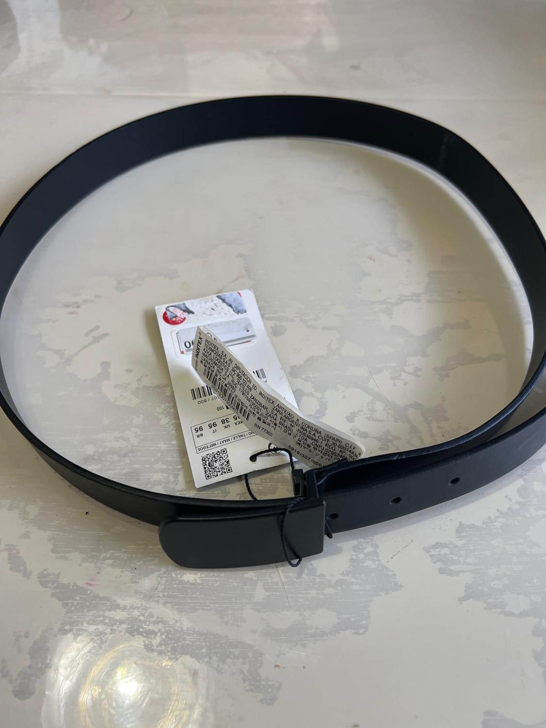 Black belt coiled in a circle with a rectangular buckle and tags. Top view on a light, distressed textured surface.