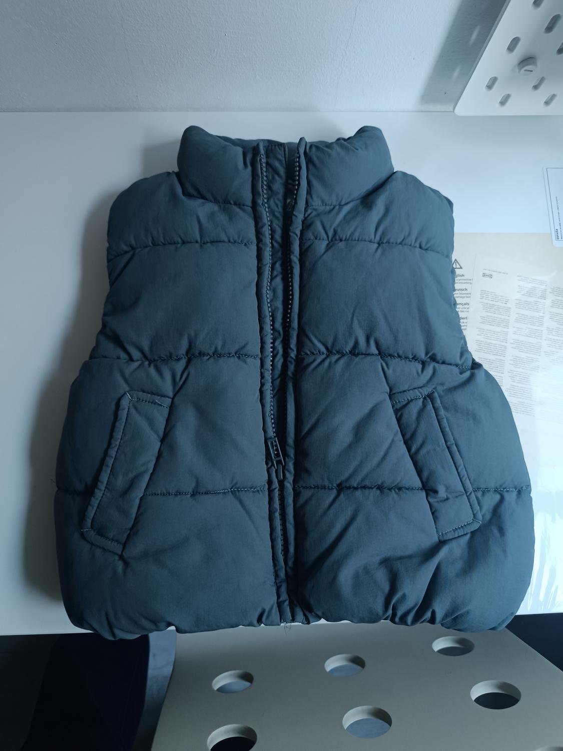 Dark grey padded gilet with front zip and pockets, on a white surface with a document background.