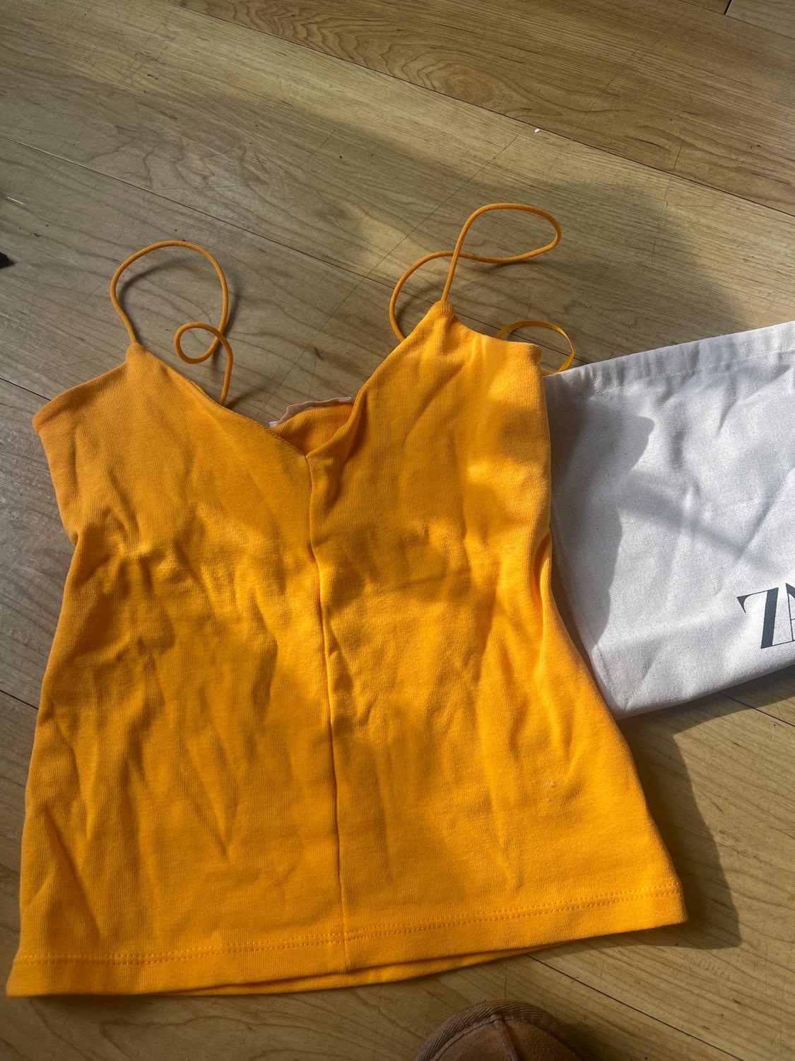 Yellow top with thin straps on a wooden floor, next to a white bag with partially visible black lettering.