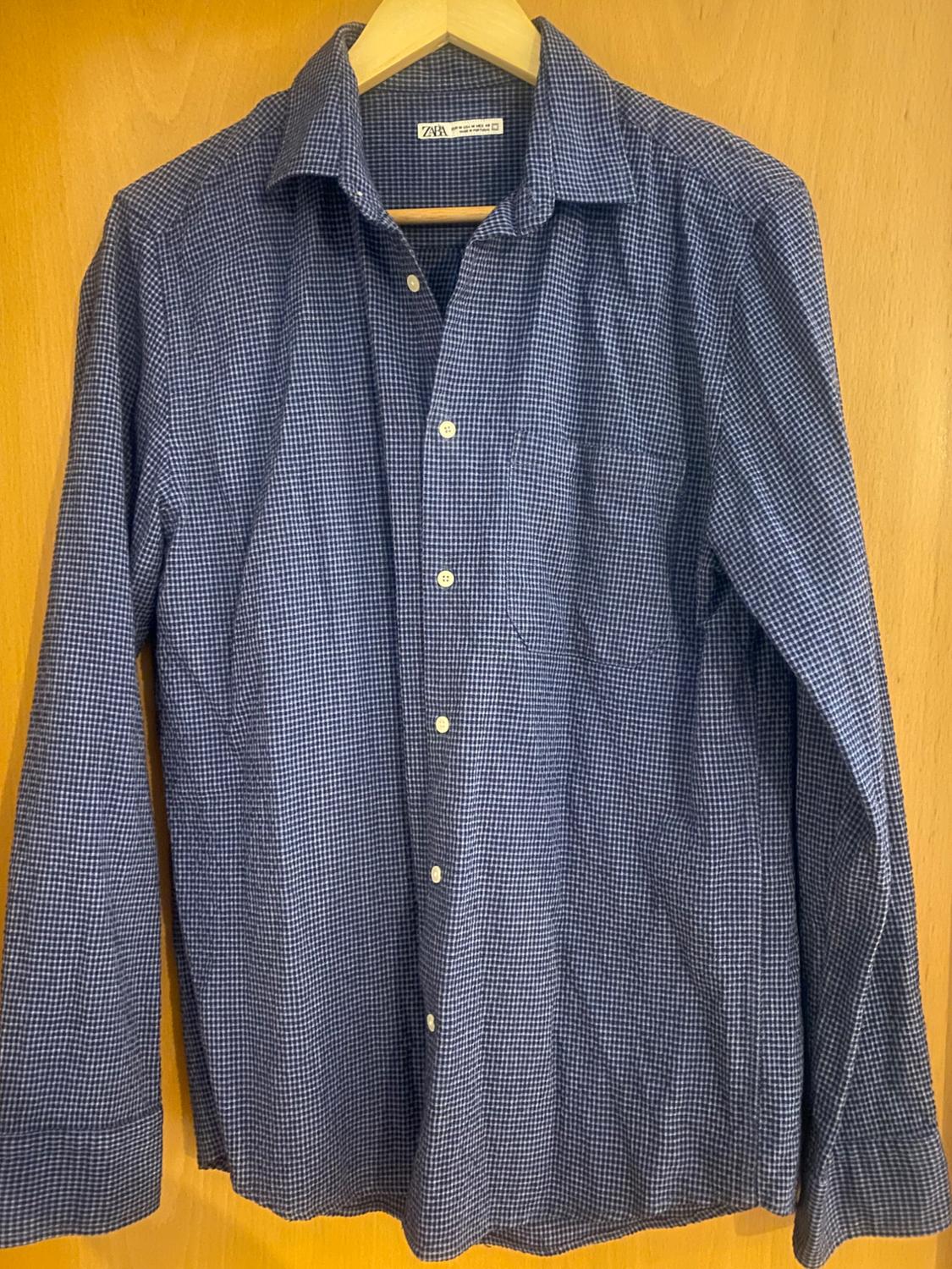Small checked shirt in dark tones hanging on a wooden hanger. Visible label with the text "ZARA". Light wooden background.