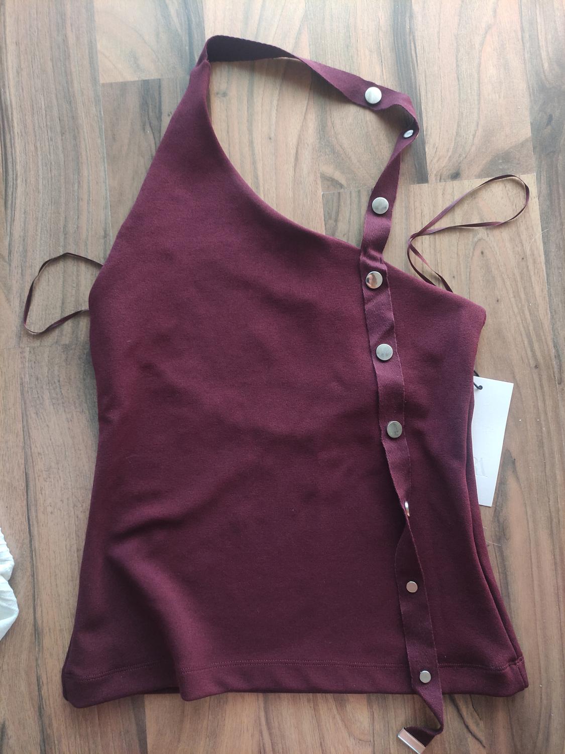Asymmetric burgundy top with straps and metal buttons on a wooden background. White label visible on one side.