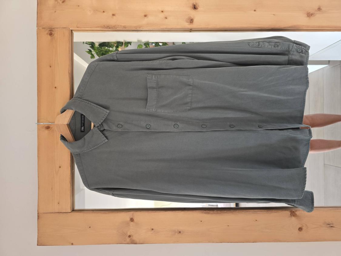 Grey-green shirt with pocket hanging in front of a wooden-framed mirror. Reflection of plants and legs. ZARA label.