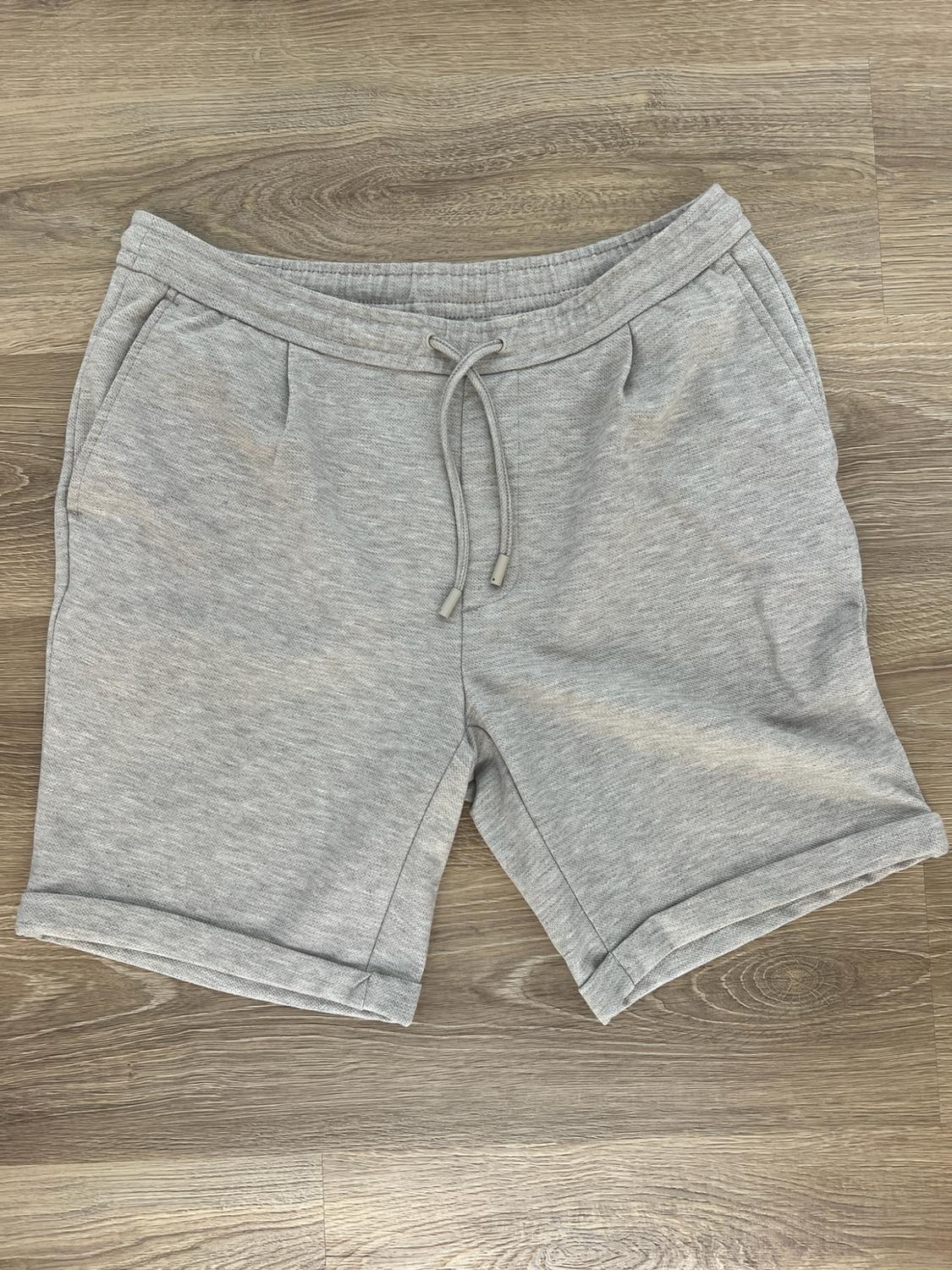 Light grey sports shorts with an elasticated waist, adjustable drawstring and turned-up hems, on a wooden floor background.