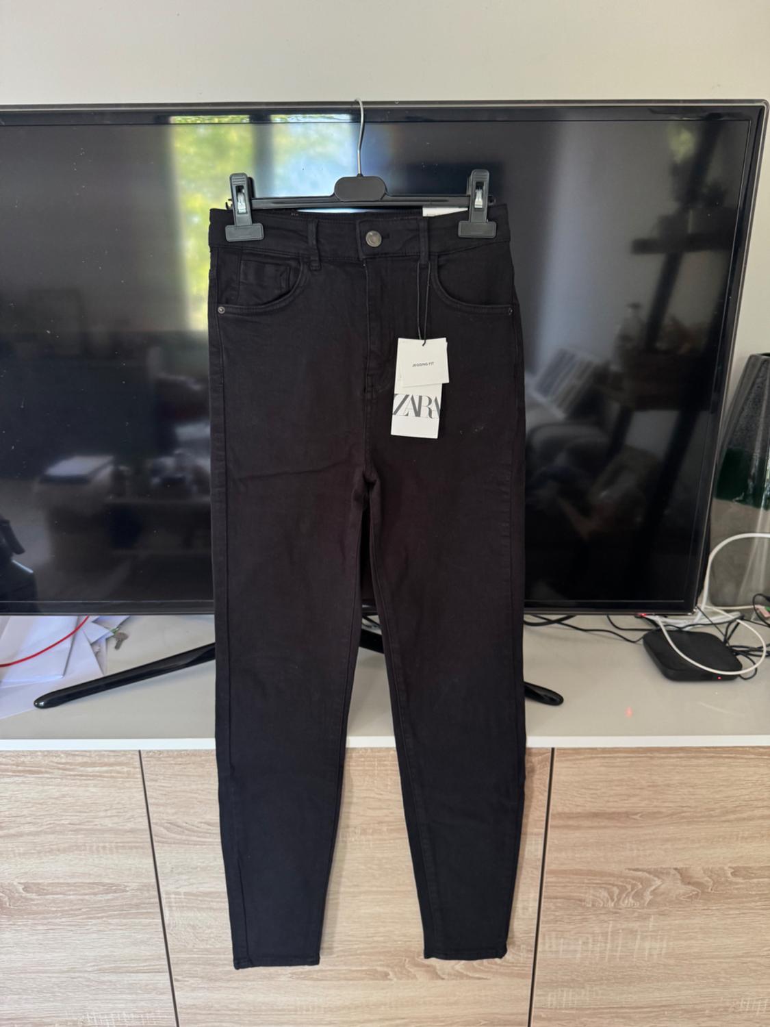 Black jeans hanging in front of a television. They have tags with the text "JEGGING FIT" and "ZARA".