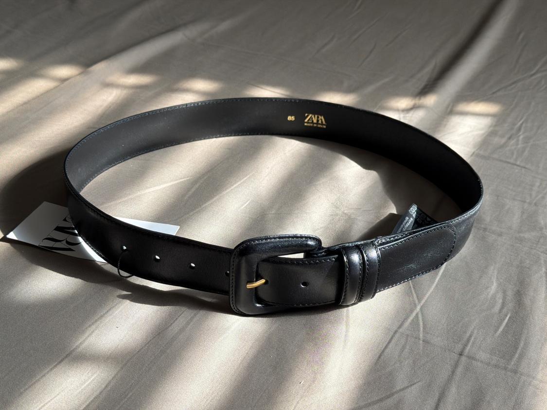 Black belt with buckle on beige fabric. Natural light creates shadows. Visible label with text "ZARA" and "Made in Spain".