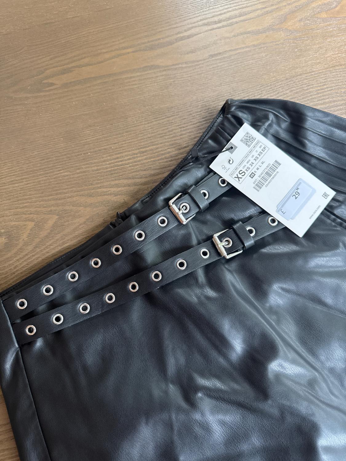 Black skirt with buckled belts and metal eyelets on a wooden surface. Visible label with size XS and price £29.99.