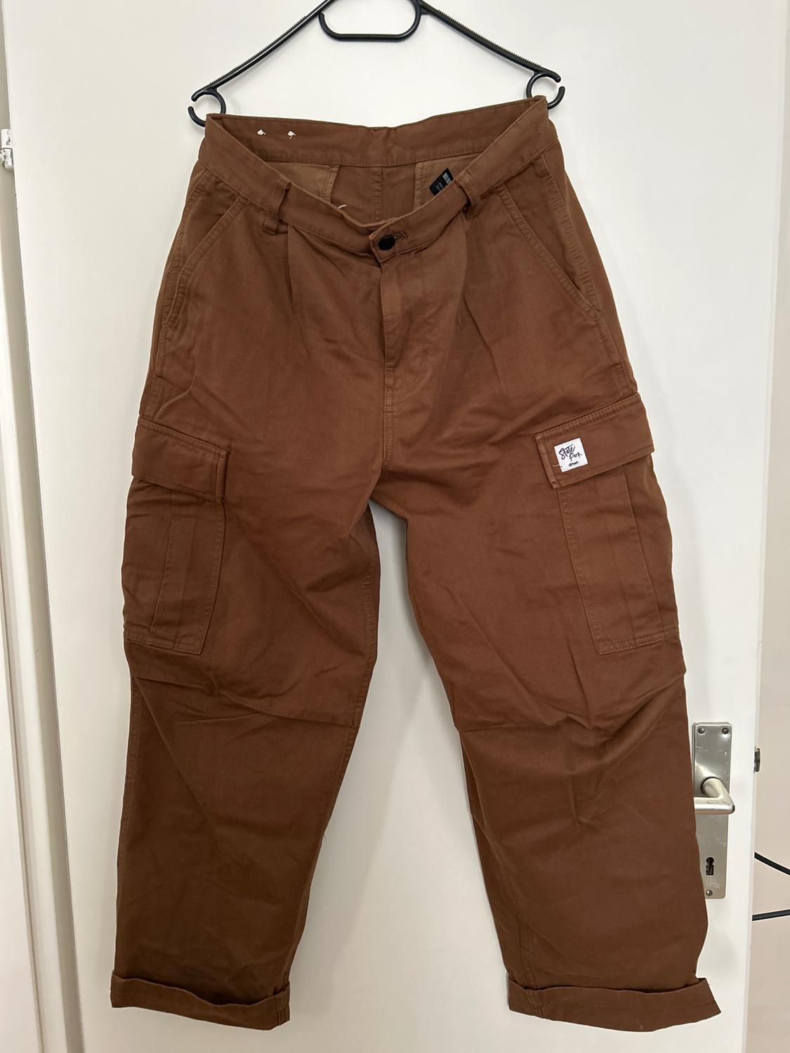 Brown trousers hanging on a black hanger over a white door. Side pockets and white label visible on the right pocket.