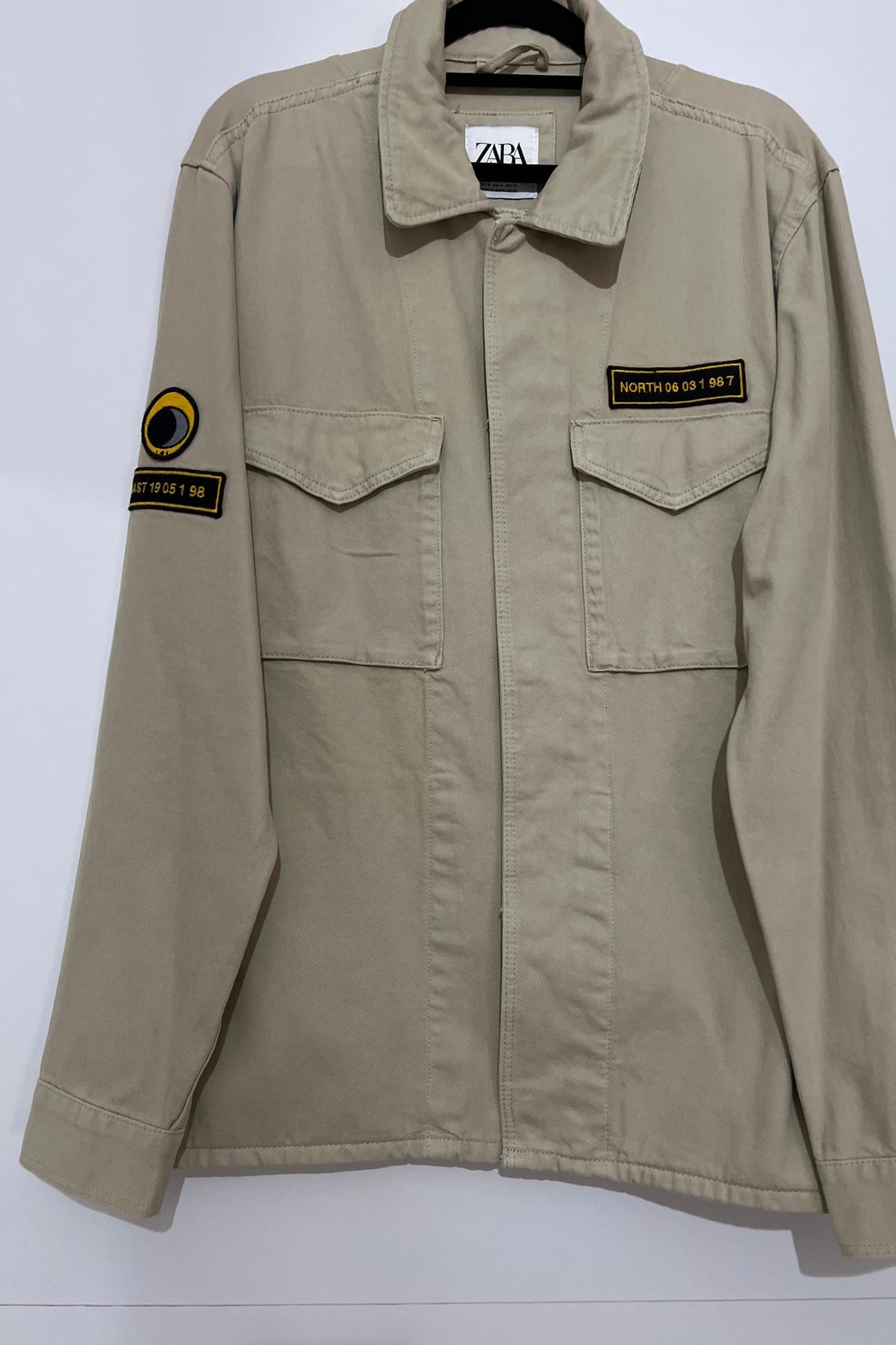 Front view of beige jacket with front pockets and embroidered patches with text 'NORTH 06 031 967' and 'EAST 19 051 98'.