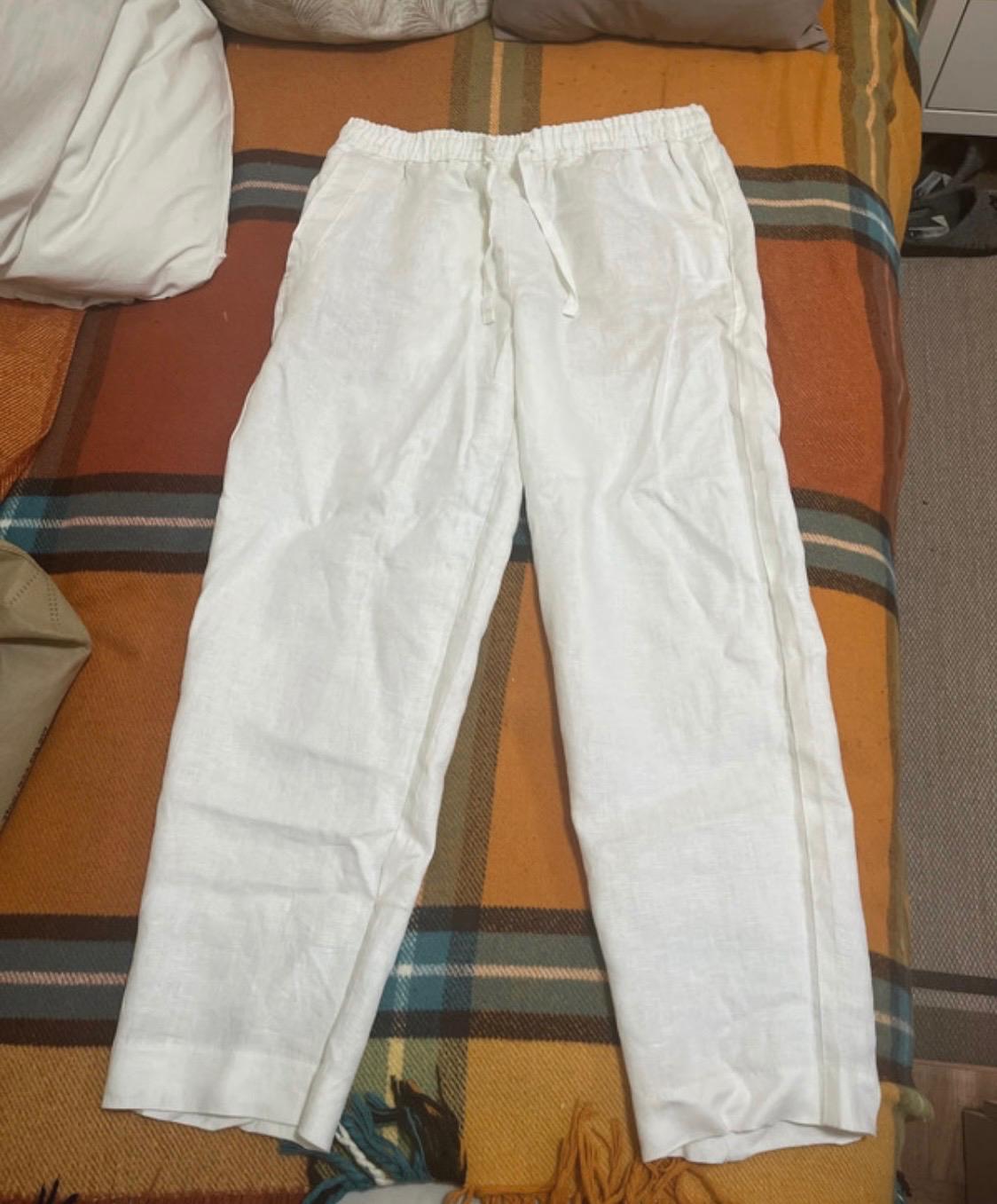 White linen trousers with an elasticated drawstring waist, on a bed with a checked blanket in shades of orange and brown.
