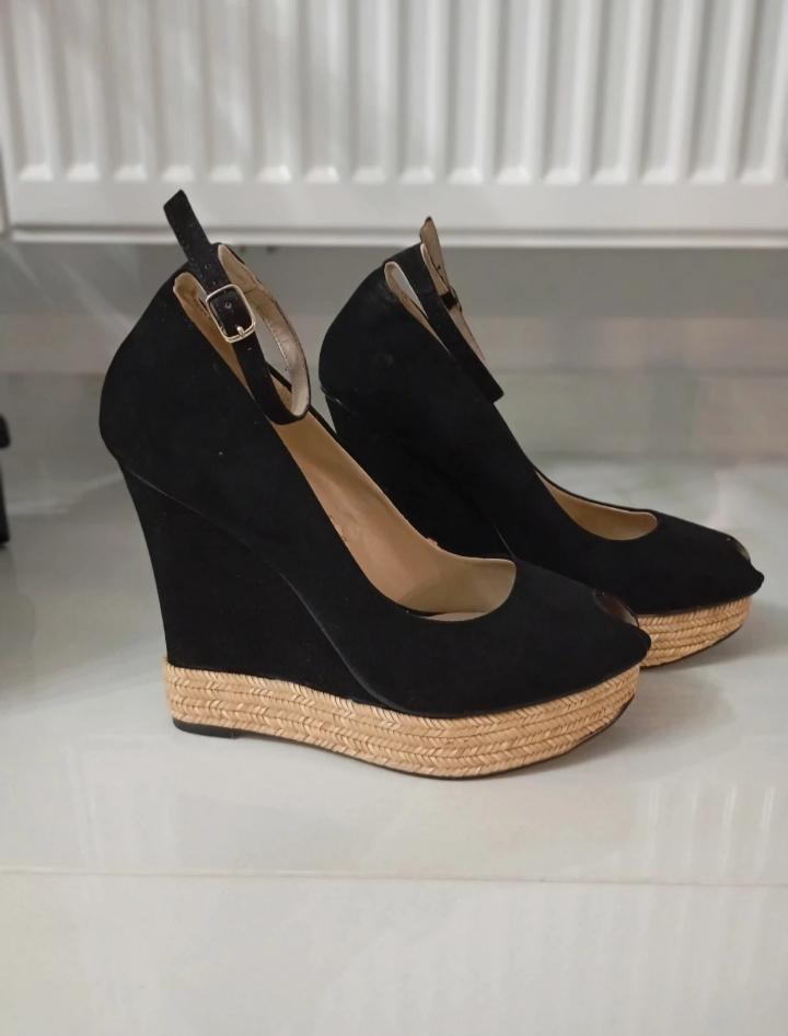 Black wedge shoes with an espadrille sole and buckle, on a shiny white floor in front of a white radiator.