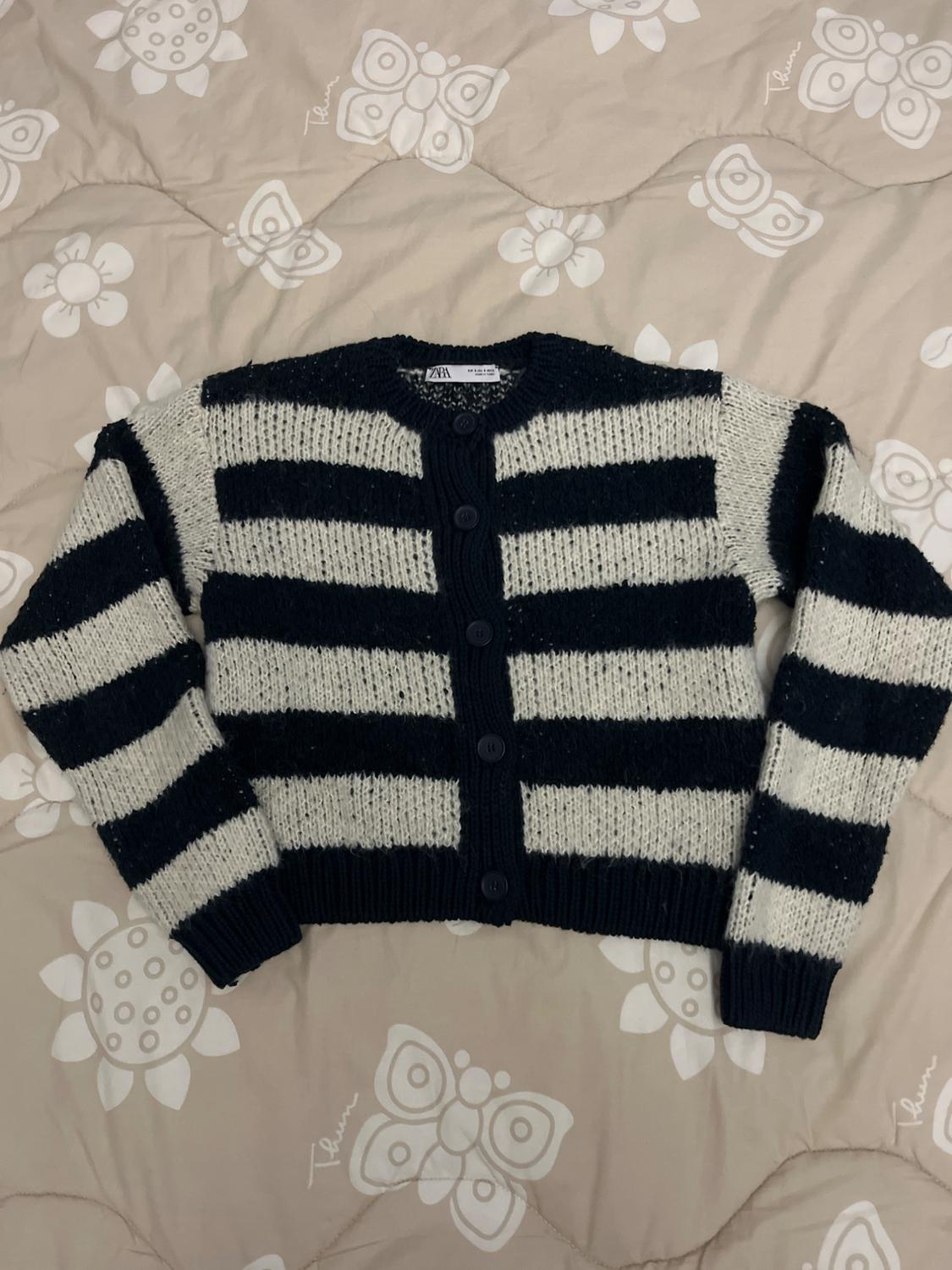 White and black striped knitted cardigan with buttons, on a beige bedspread with a butterfly and white floral print.
