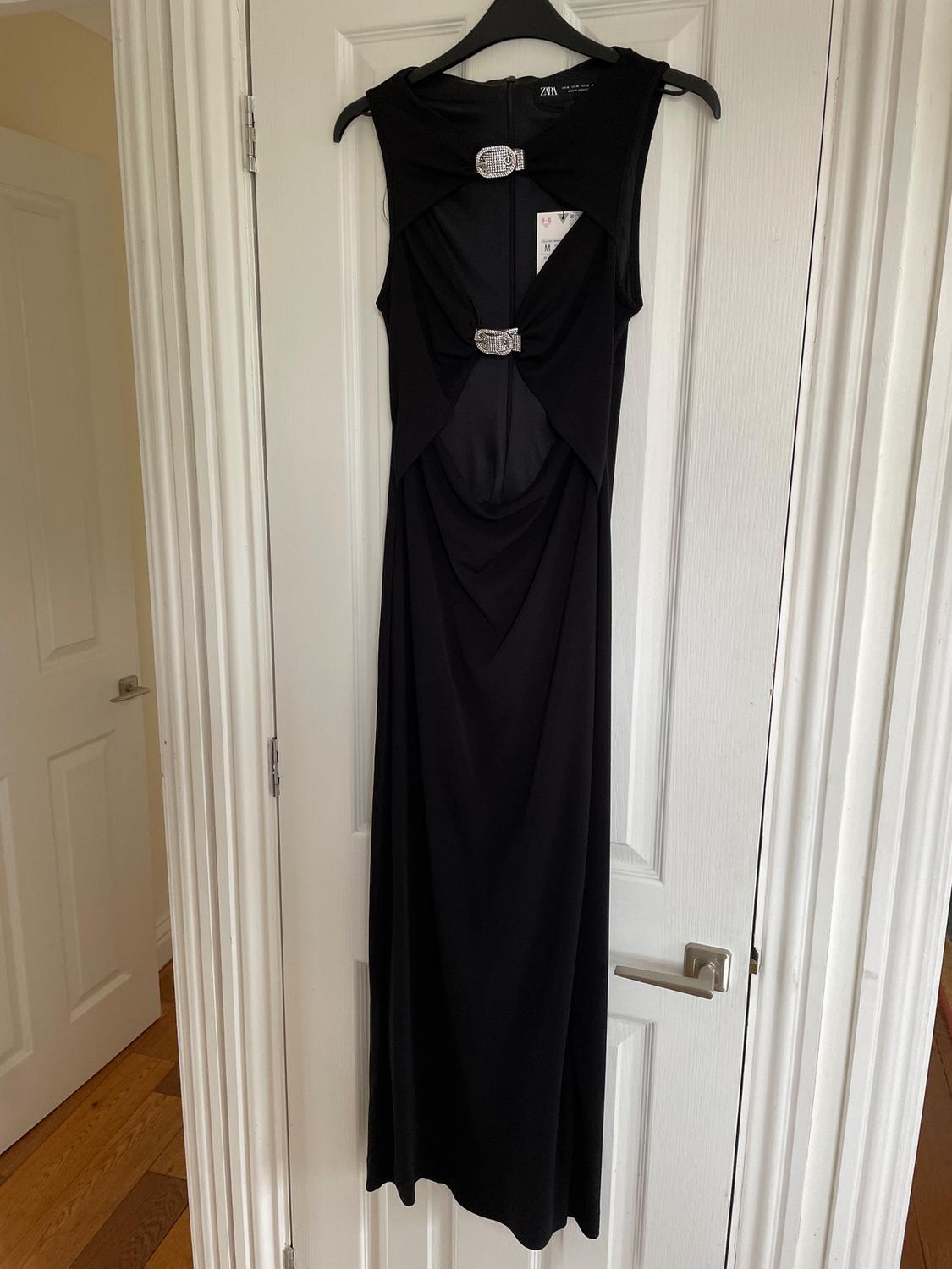 Long black dress hanging on a white door, with shiny buckle details on the chest and waist. Label visible on the inside.