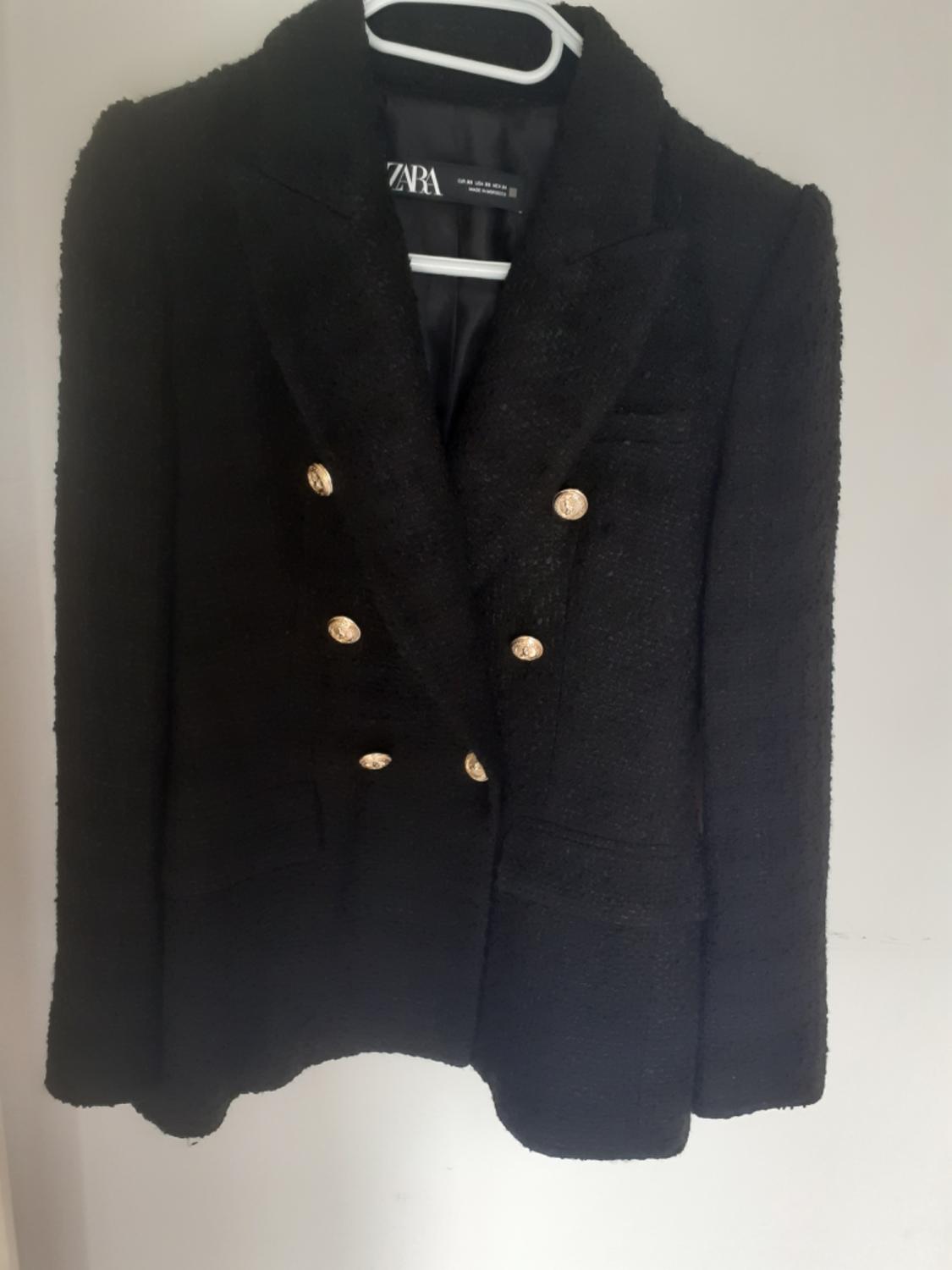 Black textured double-breasted blazer with gold buttons, hanging on a white hanger against a light background. Inside label with the text ZARA.