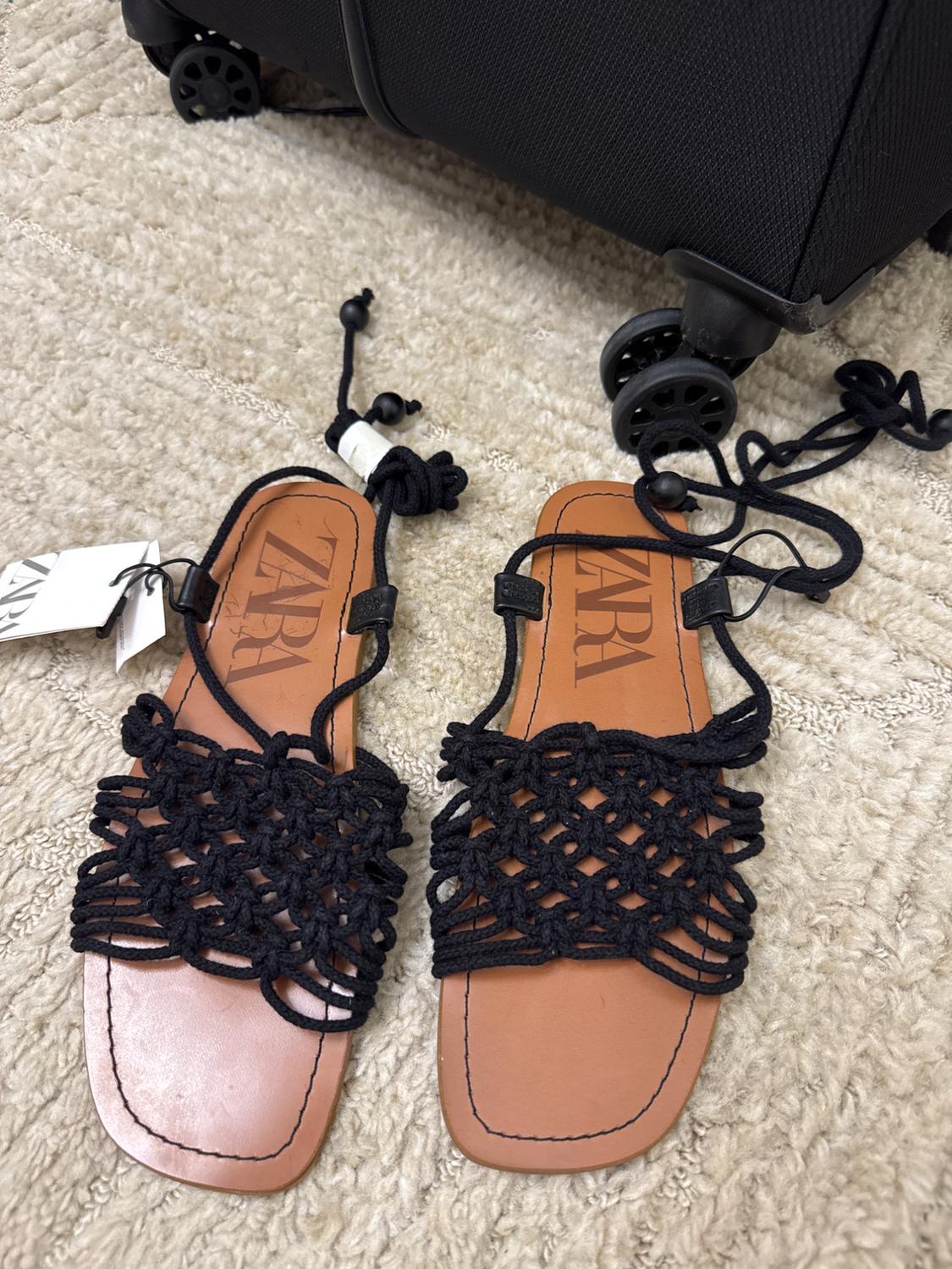 Brown leather sandals with black lace design on a beige rug, next to a black wheeled suitcase. Visible text: "ZARA".