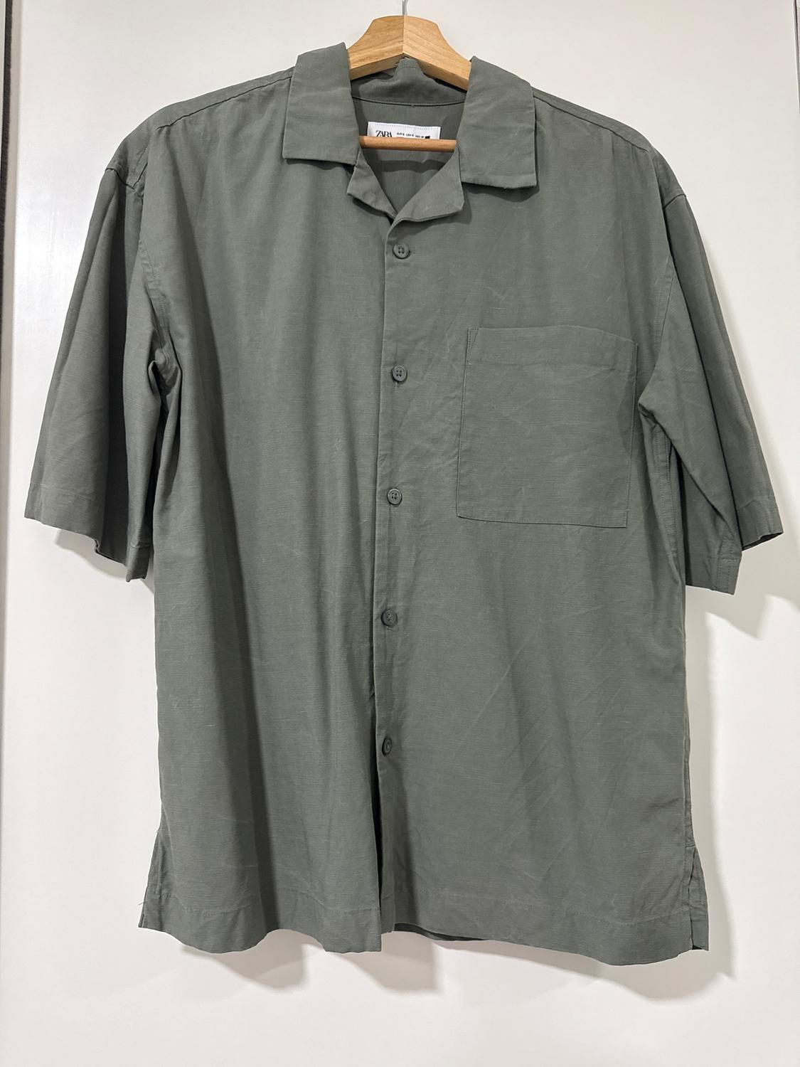 Grey short-sleeved shirt hanging on a wooden hanger, with a chest pocket and front buttons. White background.
