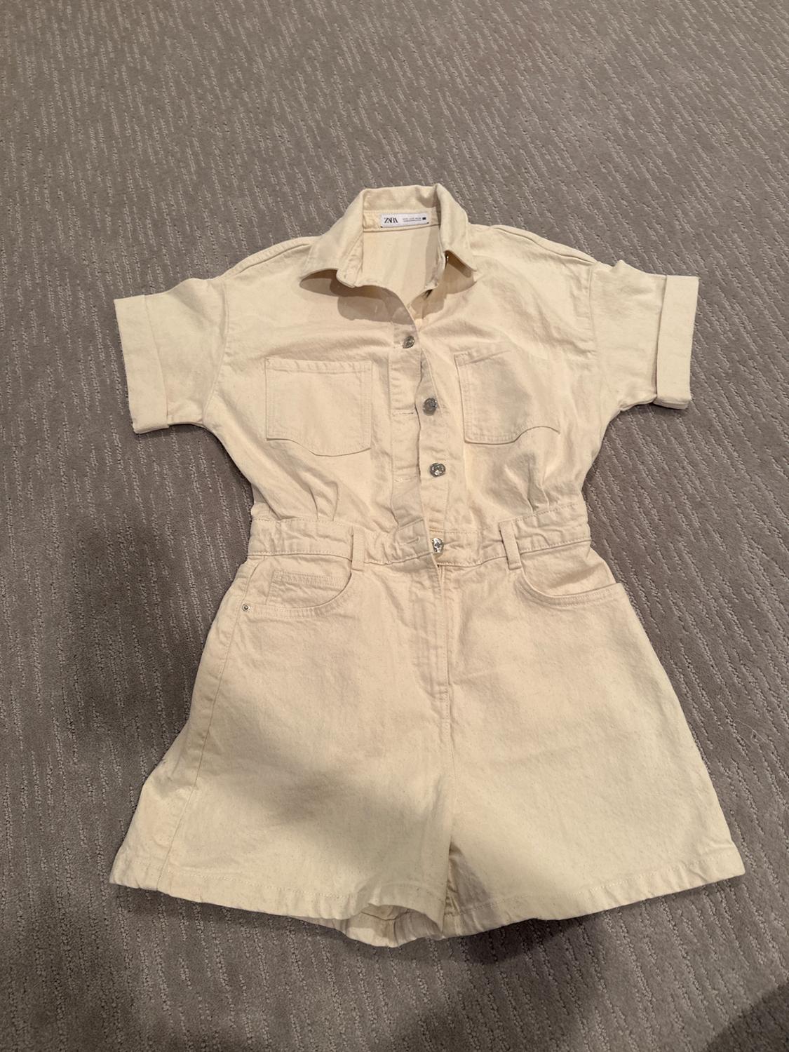 Short-sleeved beige romper with buttons and front pockets, on a gray rug. Visible tag: "Zara".
