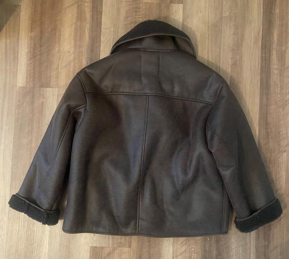 Brown leather jacket seen from behind, with black wool collar and cuffs, on a light wood floor.