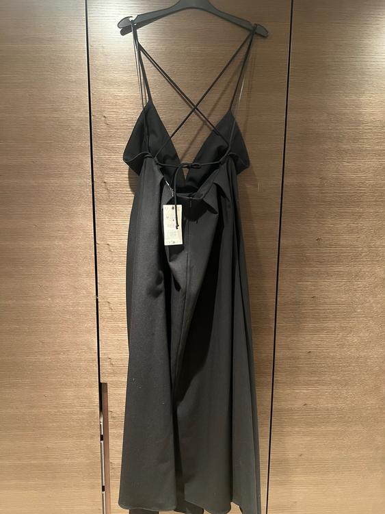 Black dress with thin cross-back straps, hanging on a hanger against a wooden background. Label visible in the centre.