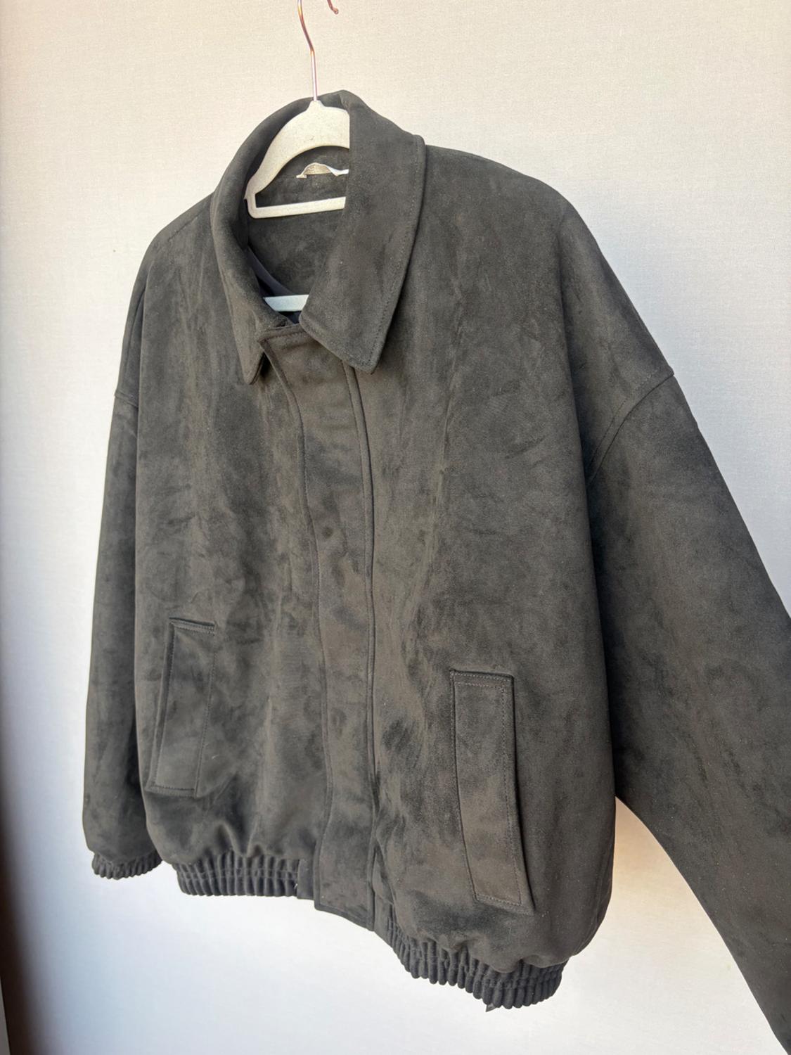 Dark grey casual jacket with a soft texture, featuring a collar, pockets, and an elasticated hem, hanging on a white hanger against a light background.