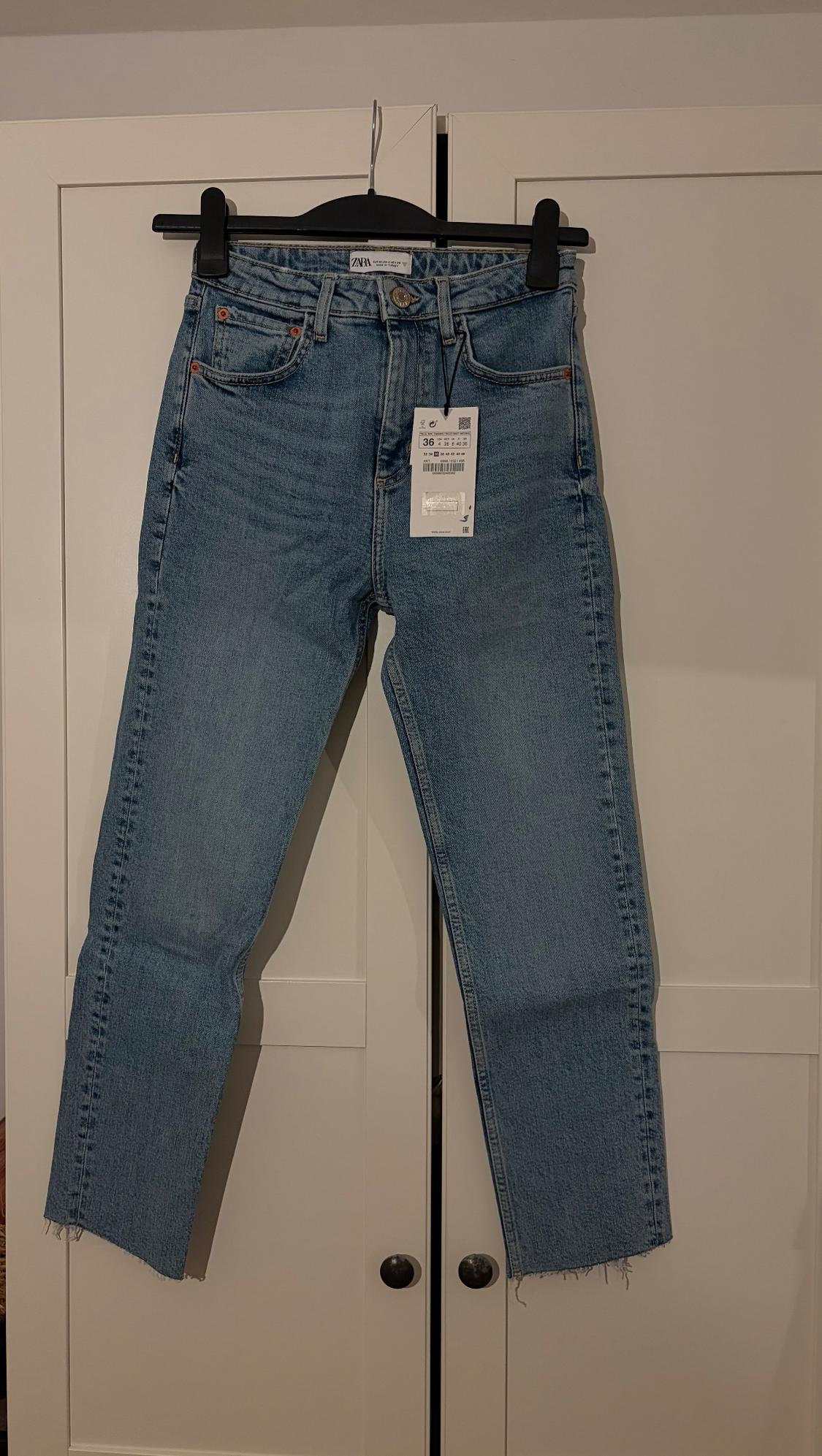 Blue jeans hanging on a black hanger in front of a white wardrobe. Visible tag with size and price details.