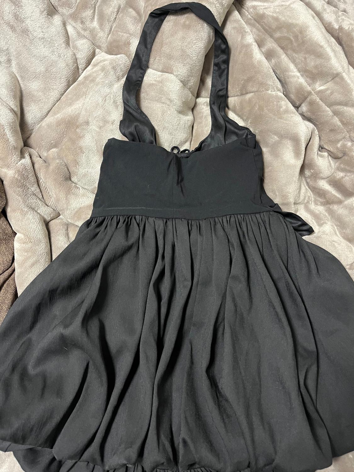 Sleeveless black dress with wide straps, on a quilted beige blanket. Soft draping fabric with pleats in the skirt.