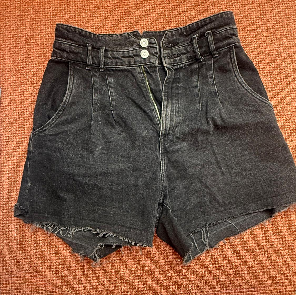 High-waisted black denim shorts with double button, open zip, darts and frayed hem on a textured orange background.