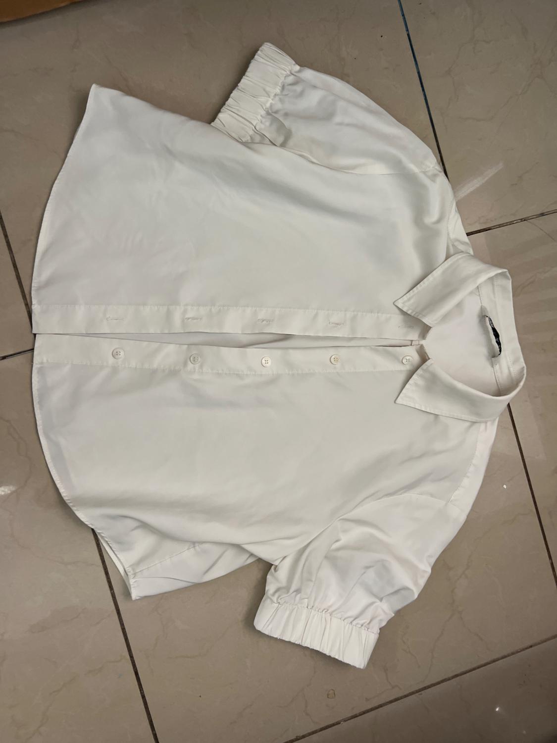 Short-sleeved white shirt with buttons and a classic collar, placed on a beige tiled floor.
