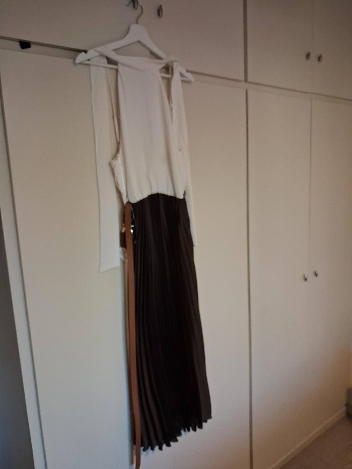 Dress hanging from a hanger in a white wardrobe. It has a sleeveless white bodice and a dark brown pleated skirt with a belt.