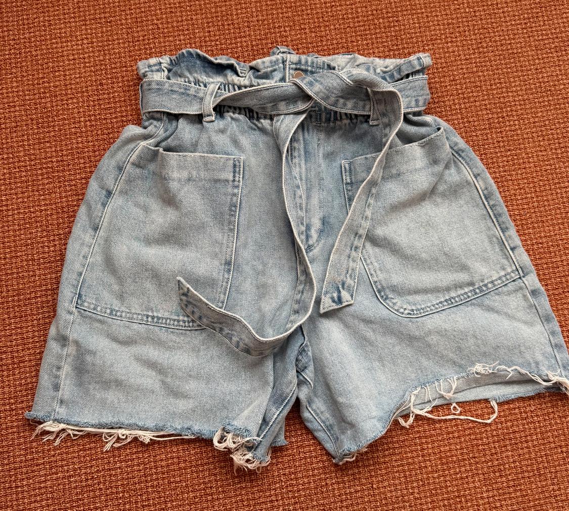 Light blue denim shorts with a gathered waist, belt, patch pockets and frayed hem on an orange background.