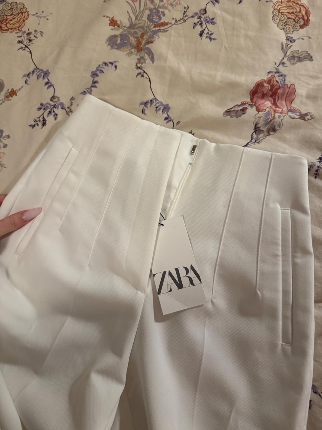 A pair of white pants with a Zara tag is on a bedspread with a floral design. A hand is holding the garment.