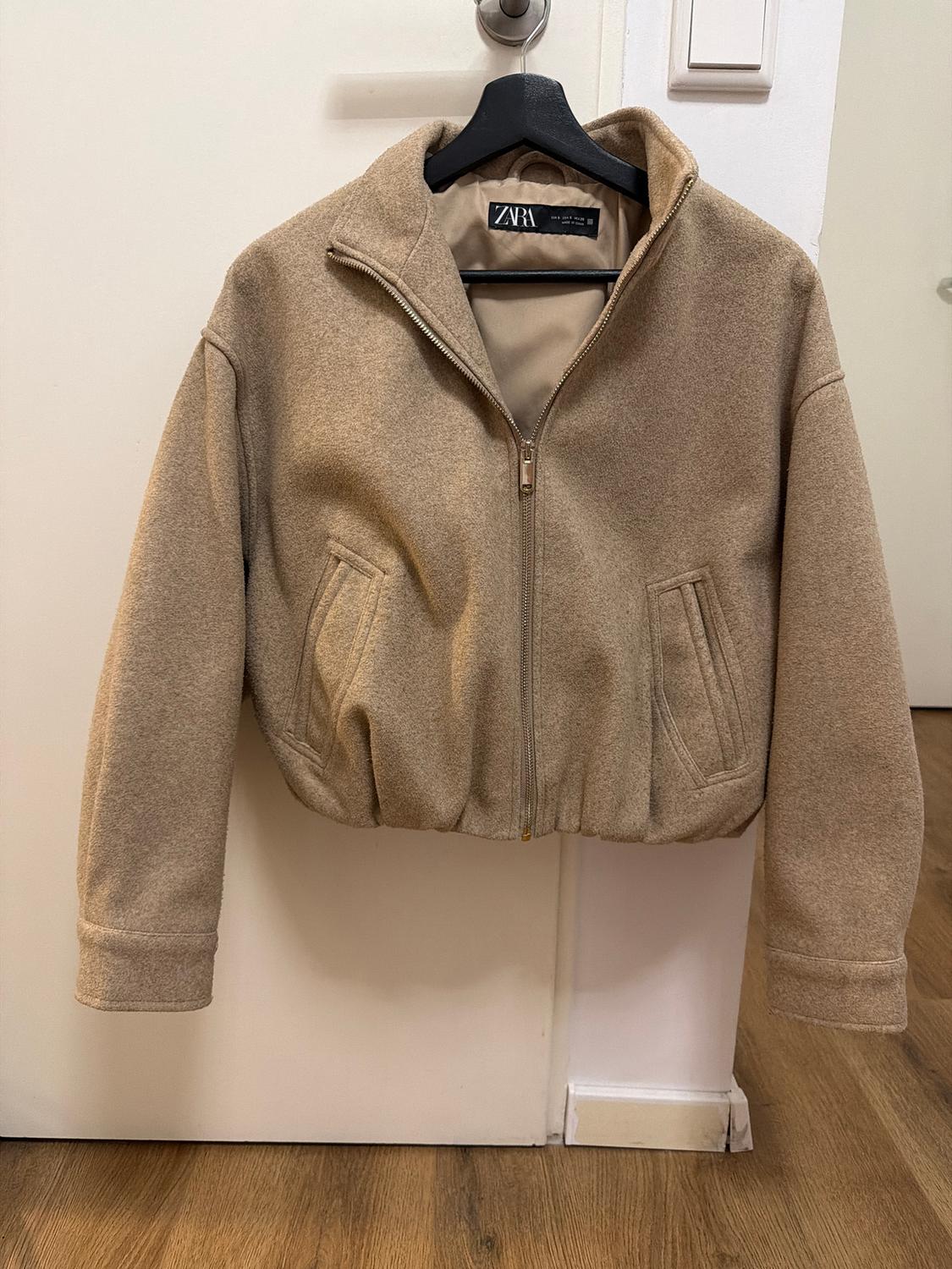 Beige jacket with zip and high collar hanging on a black hanger. Visible label with the text "ZARA". White door background.