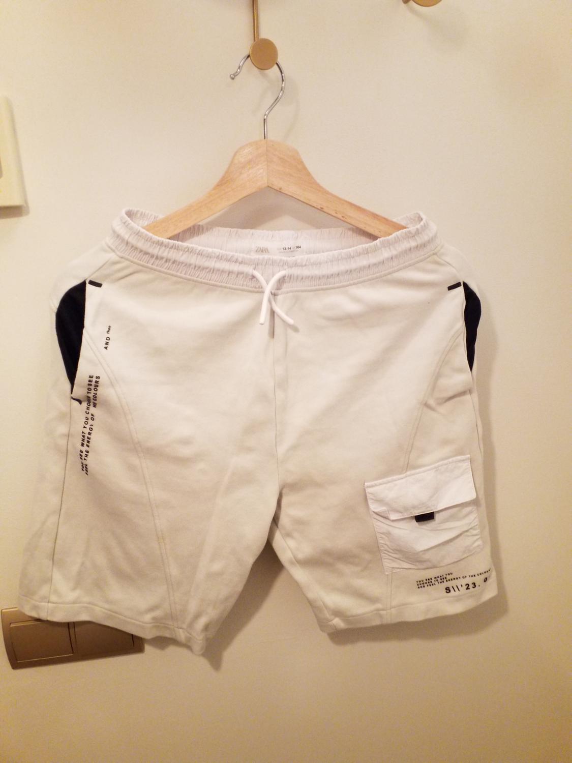 White shorts with black detailing and text on the side, hung on a wooden hanger against a white wall.