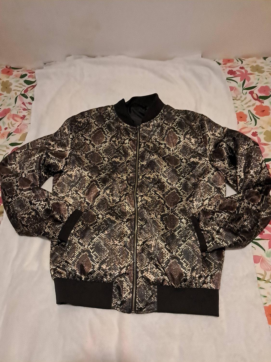 Bomber jacket with a snake print and black edging, laid out on white fabric with a floral background.