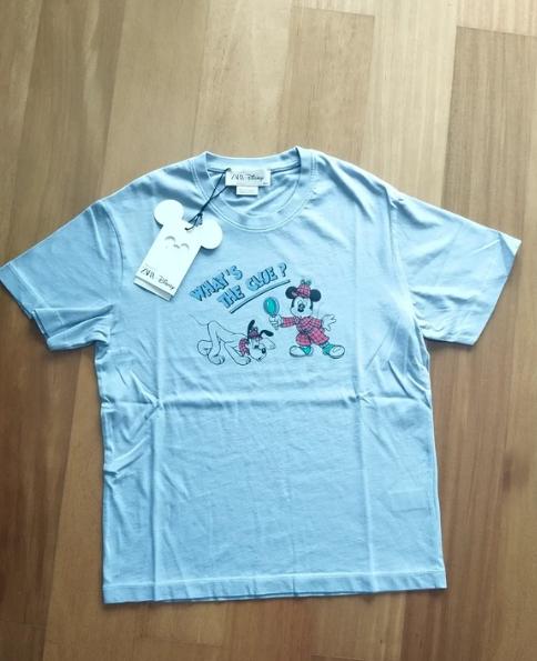 Light blue t-shirt with animated character print and "WHAT'S THE CUE?" text on the chest. Mouse-shaped hang tag.