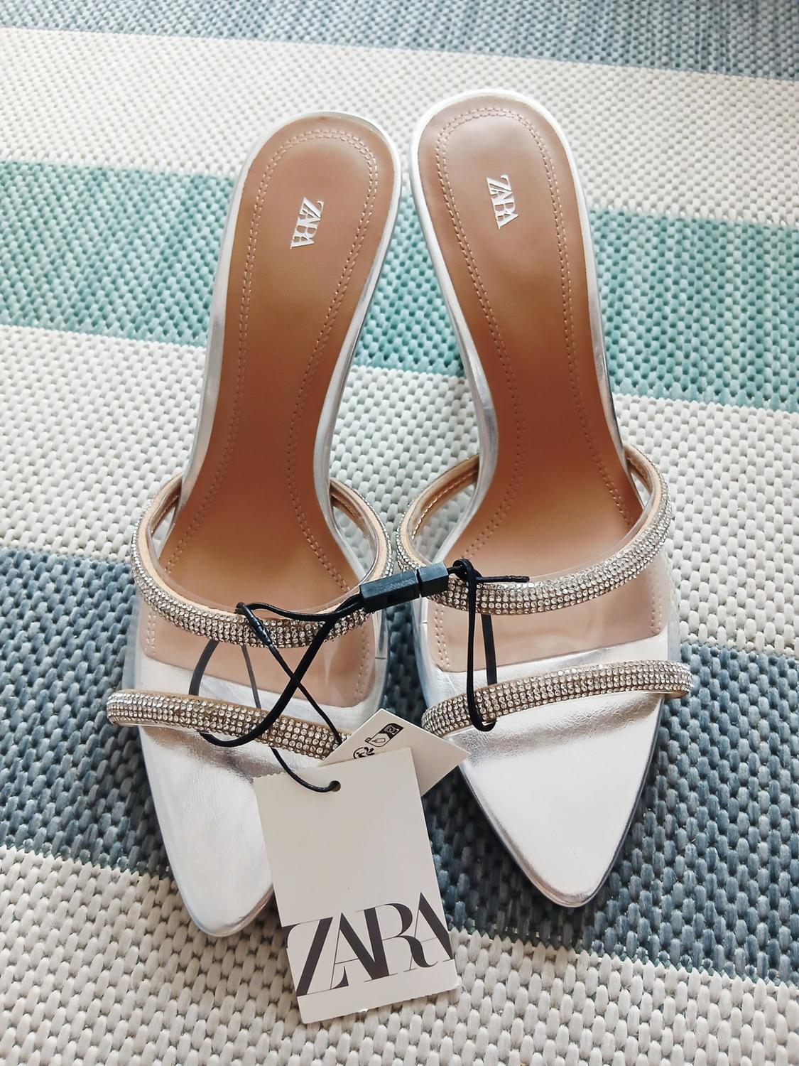 Sandalias plateadas de tacón con tiras transparentes y brillantes sobre alfombra a rayas. Etiqueta y plantillas con texto ZARA.
