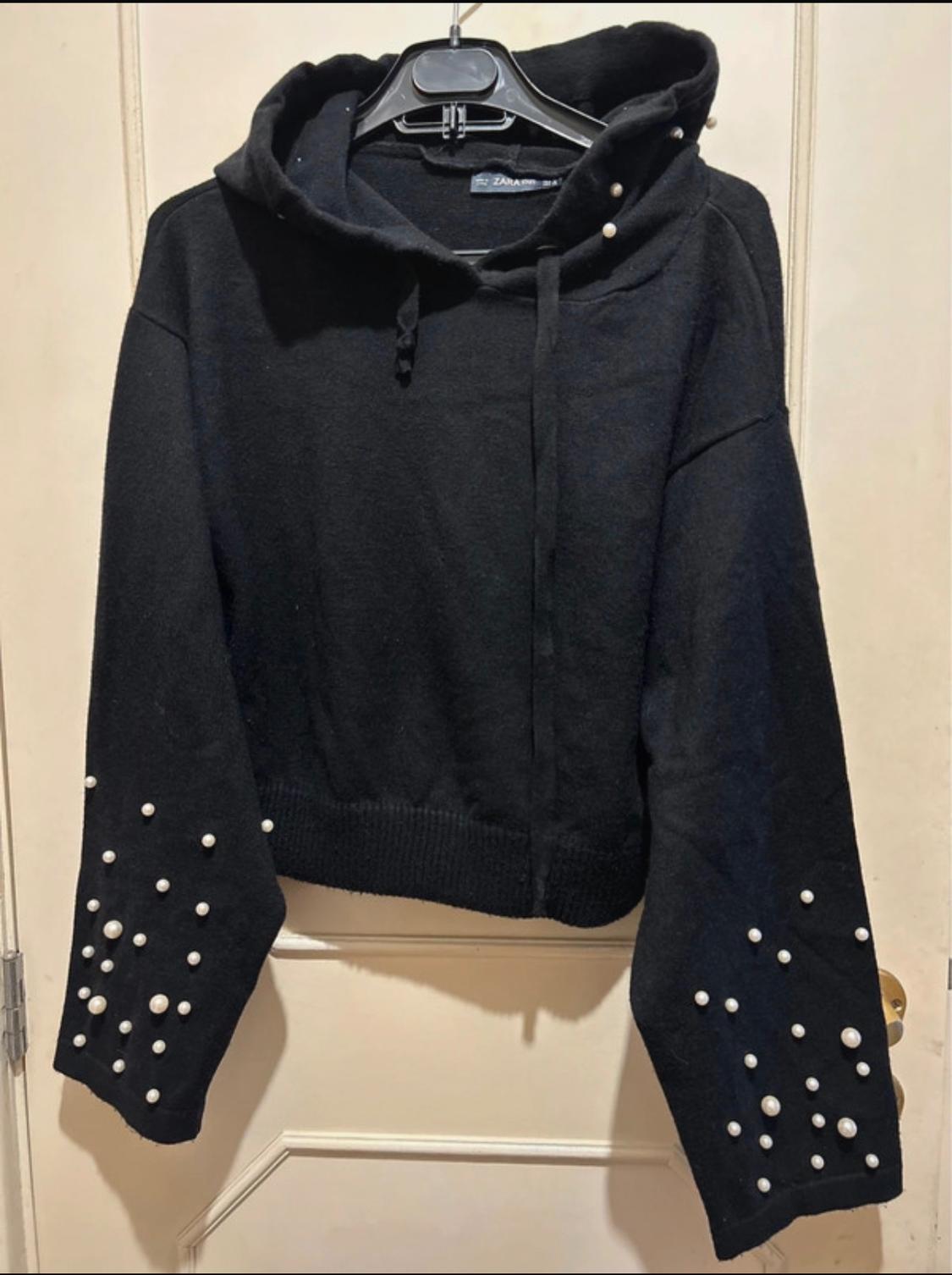 Black hooded sweatshirt with decorative pearls on the sleeves, hanging on a black hanger in front of a white door.