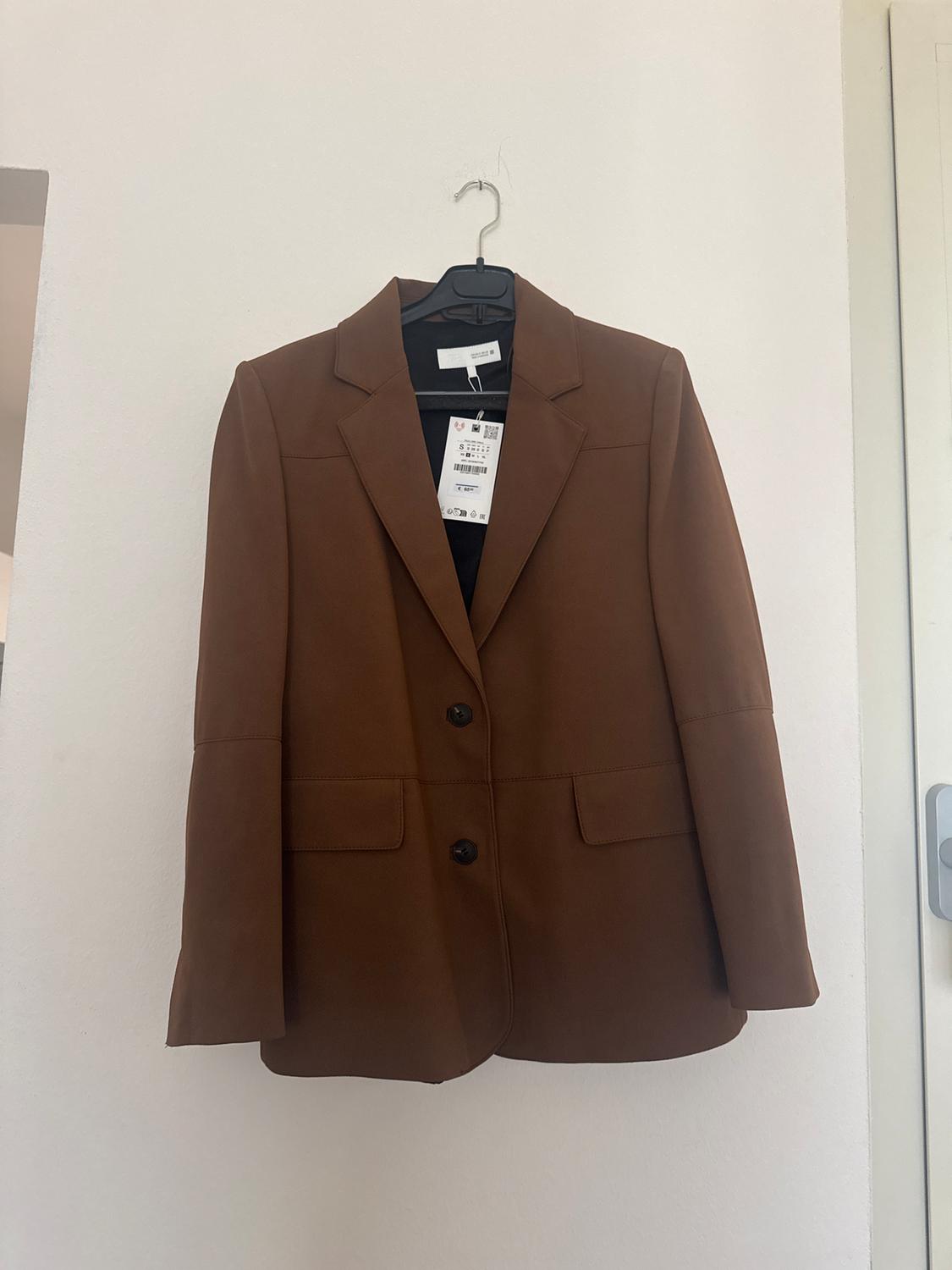 Brown jacket hanging on a black hanger against a white wall. Visible label with barcode and text.