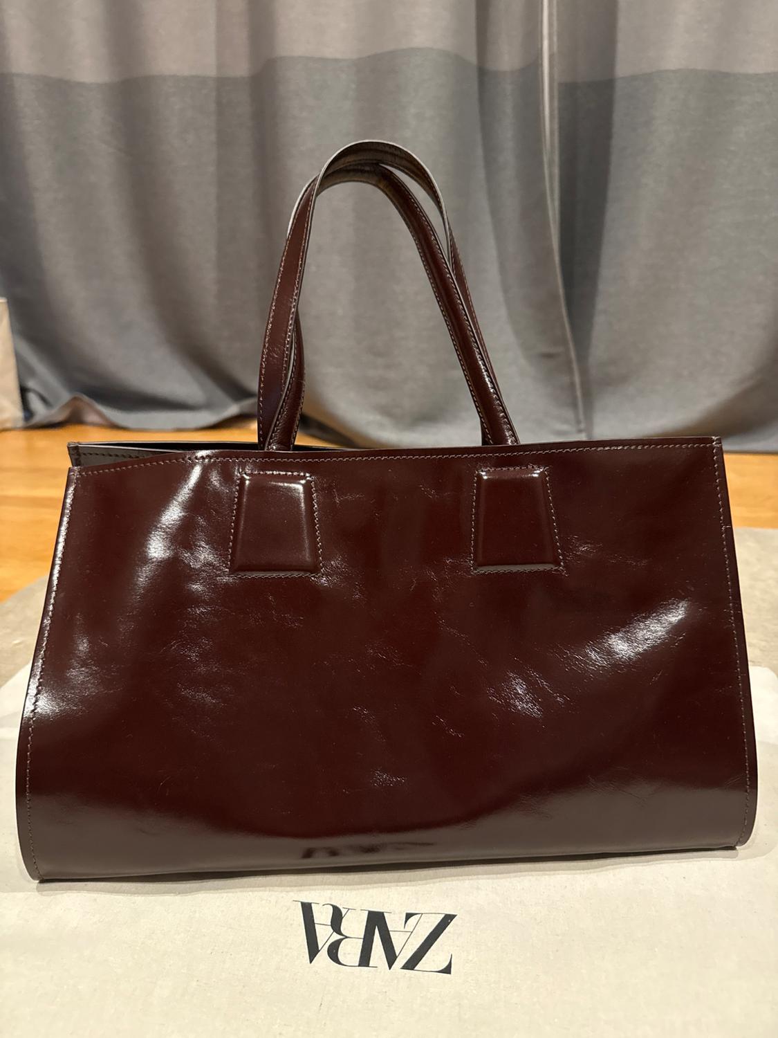 Shiny burgundy tote bag on white fabric with the text ZARA. Background with wooden floor and grey curtain.