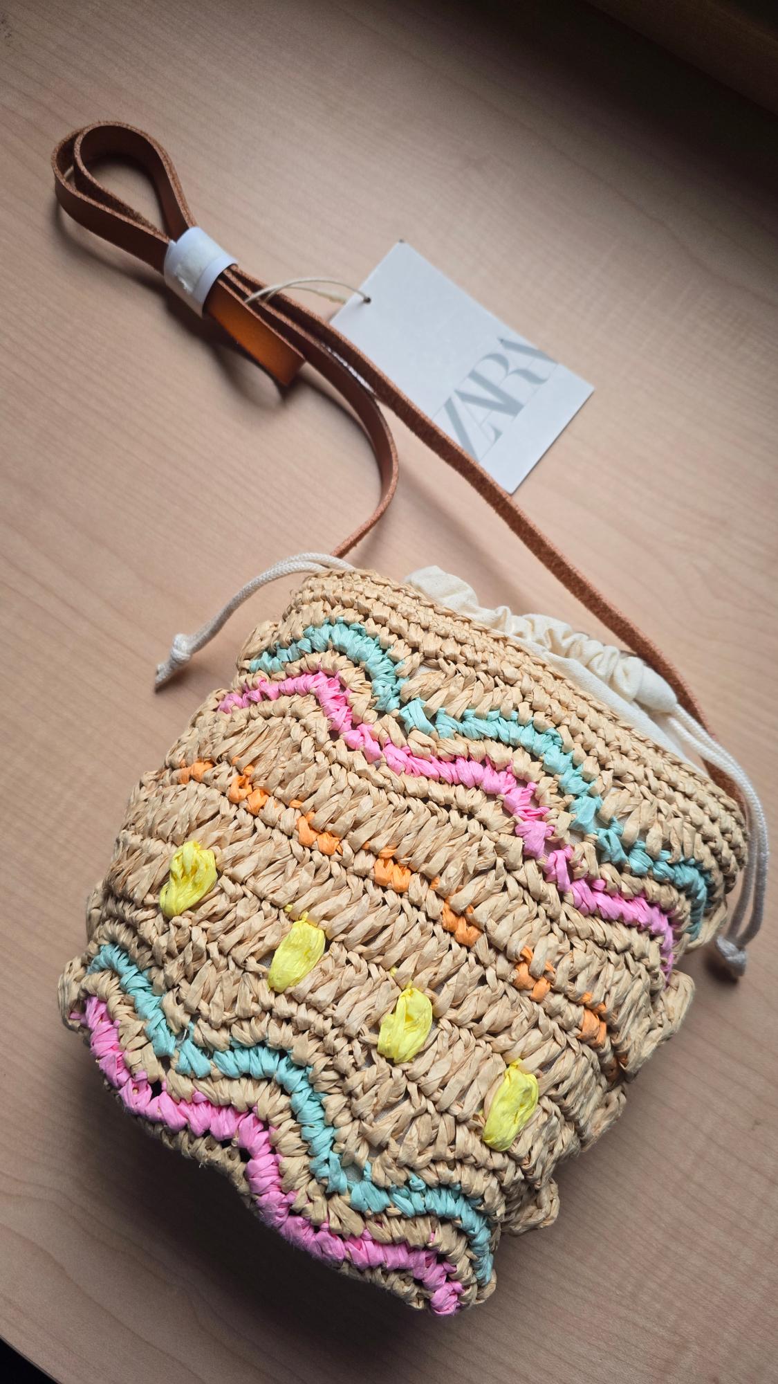 Woven handbag with a pastel wave pattern on a wooden table. Brown leather strap and visible label with "ZARA" text.