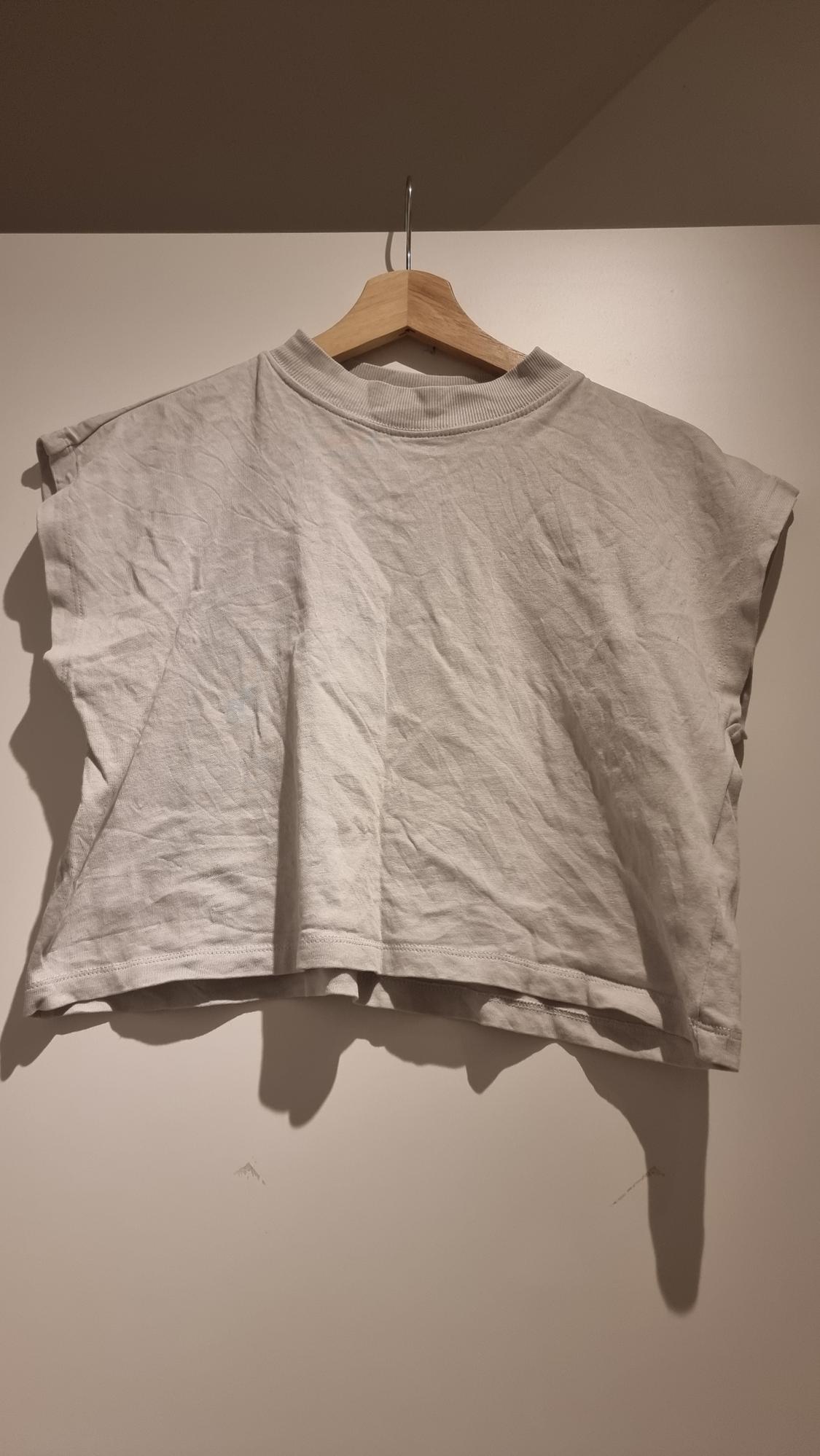 White sleeveless crop top hanging on a wooden hanger against a white background. The fabric has a wrinkled appearance.