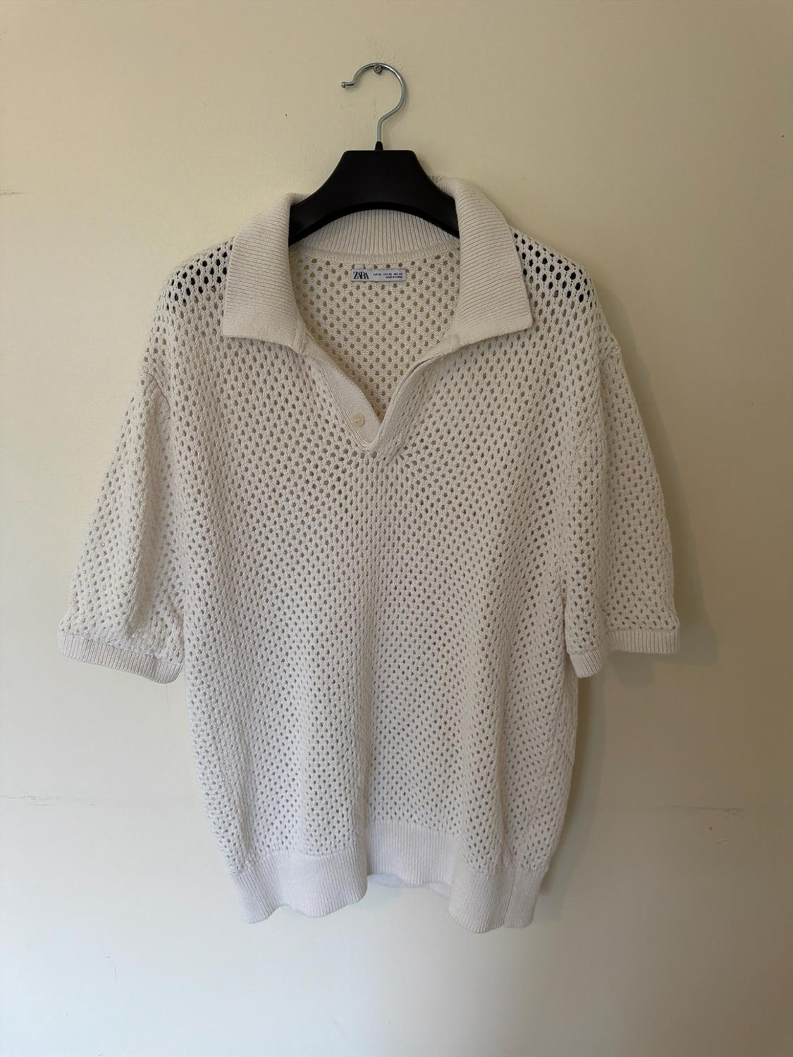 White open-knit polo shirt with a collar and short sleeves, hanging on a black hanger against a light background. Interior label with ZARA text.