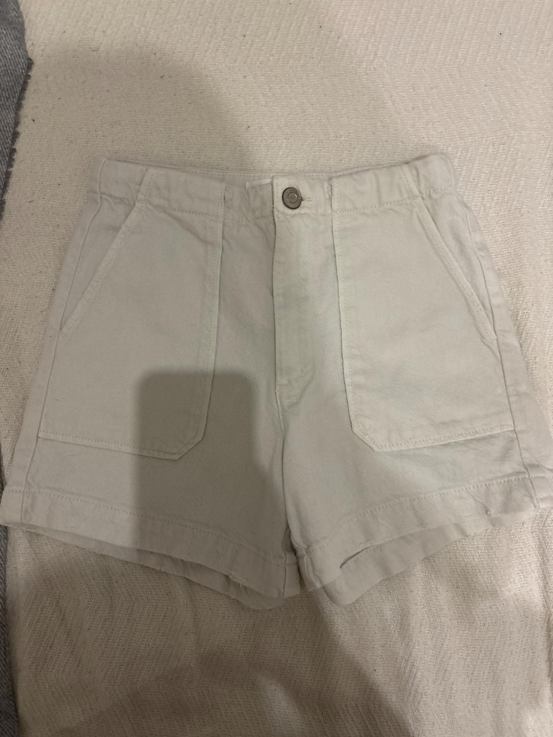 Beige shorts with front pockets on a light textured surface. Metal button at the waist.