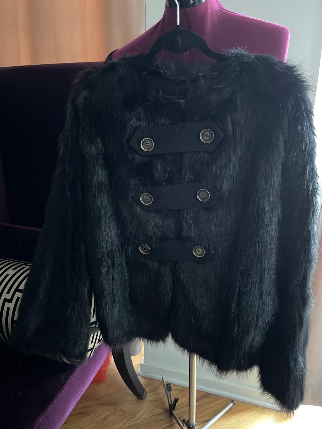 Black faux fur coat with large buttons, hanging on a purple mannequin. Background with chair and geometric design cushion.