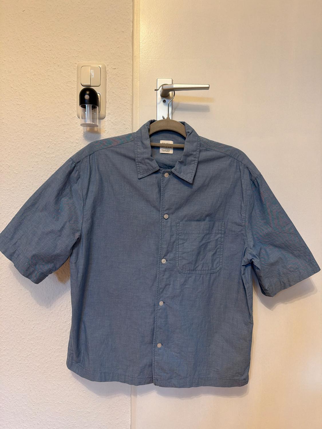 Blue short-sleeved shirt hanging on a white door. Beige wall background with visible soap dispenser and plug socket.