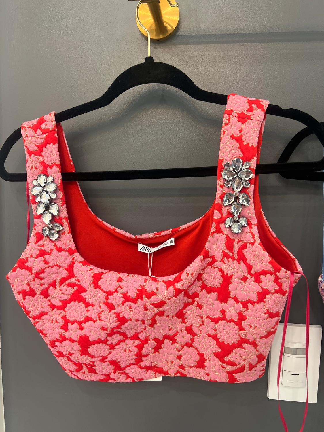 Red crop top with pink floral print and sparkly embellishments on the straps, hanging on a black hanger against a gray wall.